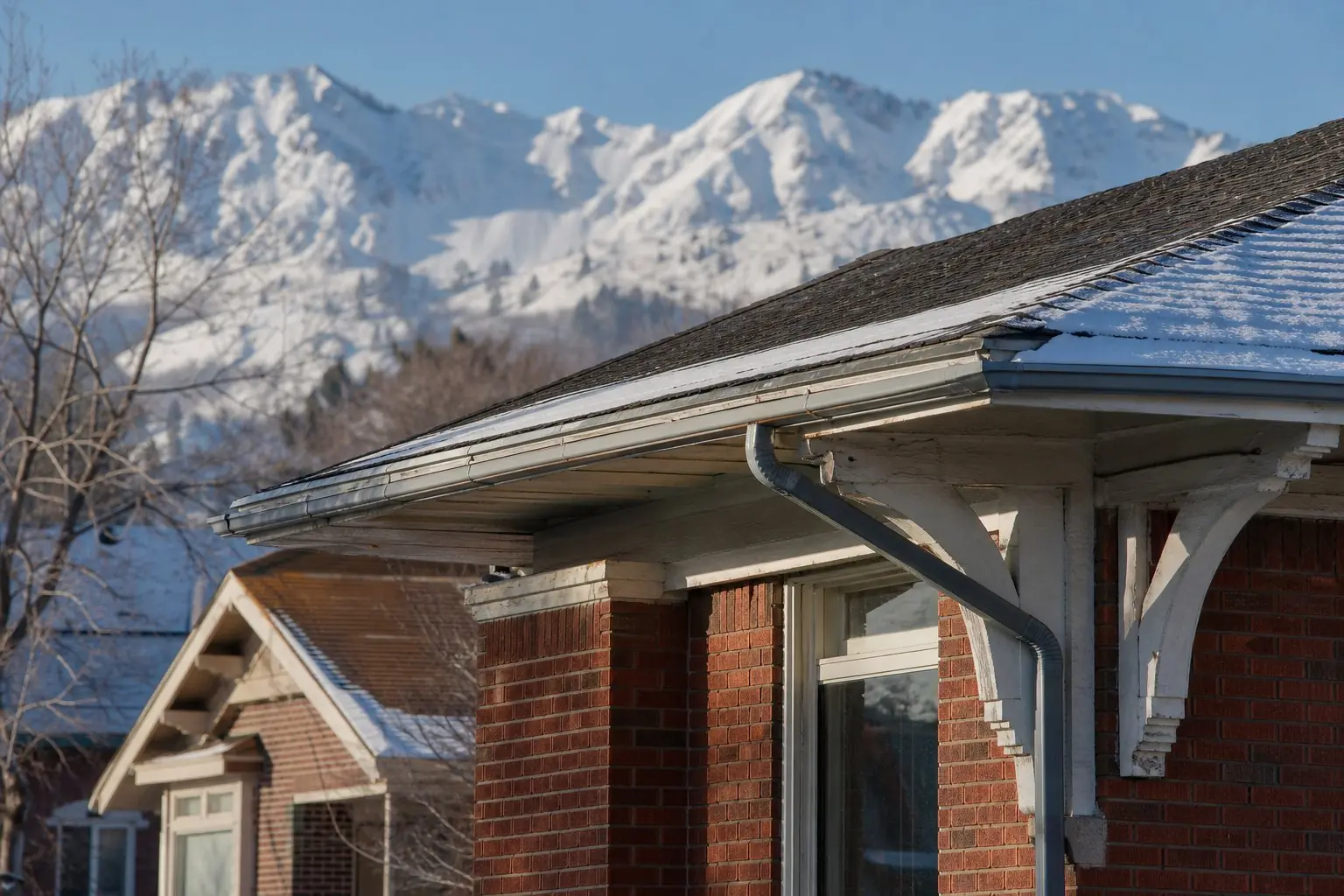 The roof edge, gutters and painted trim of an older brick home with snowy mountains behind